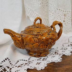Op de afbeelding: Een bruine theepot van keramiek met een gedetailleerd ontwerp van druiven en bladeren. De theepot heeft een rond lichaam en een gebogen tuit.