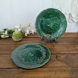 May include: Two round ceramic plates with a deep green glaze and a textured leaf pattern. One plate rests flat while the other is displayed upright on a small metal stand. The surface of each plate features a detailed embossed foliage design.