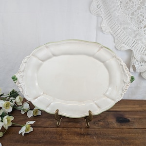 Op de afbeelding: Een crèmekleurige ovale serveerschaal met een geschulpte rand en decoratieve details. De schaal heeft groene accenten en wordt op een messing standaard gepresenteerd. Een bloemstuk en een kanten kleedje zijn op de achtergrond.