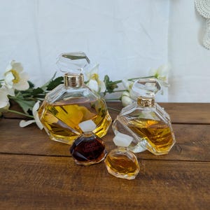 Puede incluir: Cuatro frascos de perfume de diferentes tamaños se exhiben sobre una superficie de madera. Los frascos tienen cuerpos de vidrio transparente facetado y detalles dorados. El frasco más grande tiene un tapón transparente y geométrico. El frasco más pequeño tiene el texto "Gianni Versace".