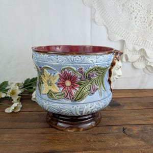 Może przedstawiać: Wazon ceramiczny w stylu vintage z jasnoniebieskim wykończeniem, z wytłoczonymi wzorami kwiatowymi w kolorze żółtym, czerwonym i zielonym. Obręcz jest bordowa, a rzeźbiona twarz zdobi bok. Wazon stoi na brązowej podstawie.