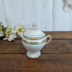 May include: A white porcelain sugar bowl with a lid, featuring gold and teal green bands. The bowl has a small handle and sits on a pedestal base. The lid is topped with a decorative finial. The sugar bowl is on a wooden surface.