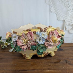 May include: A cream-colored ceramic planter with a scalloped rim, adorned with sculpted pink, blue, and yellow flowers and green leaves. The planter sits on a wooden surface, showcasing a vintage floral design.
