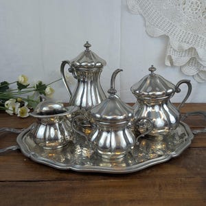 Op de afbeelding: Een zilveren theeservies, bestaande uit een theepot, een melkkan, een suikerpot en een serveerblad. De set heeft een gegroefd ontwerp en wordt gepresenteerd op een houten oppervlak. De lade heeft handvatten. De achtergrond omvat witte bloemen en een kanten kleedje.