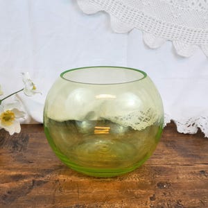 May include: A round, green glass vase with a slightly iridescent finish. The vase is sitting on a wooden surface.