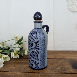Op de afbeelding: Een steengoed fles met een lichtgrijze behuizing, diepblauwe reliëf bloemmotieven en een verticale textuurband. De fles is voorzien van een handvat aan de zijkant, een grijze hals en een afgeronde donkerblauwe stop met een kurken basis.