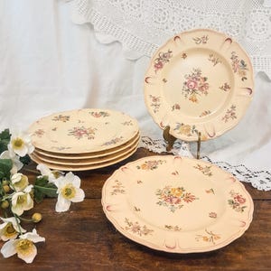 Può includere: Set di sei piatti da portata in ceramica color crema con un motivo floreale. I piatti hanno un bordo ondulato e sono decorati con fiori rosa, gialli e verdi.