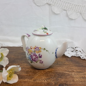 Peut inclure: Une théière en porcelaine blanche avec un corps rond et un couvercle. La théière est décorée de motifs floraux colorés violets, jaunes et bleus. La poignée et le bec ont des accents violets et bleus. La théière est posée sur une surface en bois.