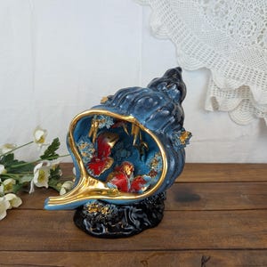 Op de afbeelding: Een decoratief keramisch sculptuur in de vorm van een schelp met een diepblauwe buitenkant en een glanzende, goudkleurige rand. Binnenin de schelp bevinden zich drie kleine rode visfiguren tussen gestructureerde blauwe en gouden koraalaccenten.