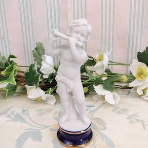 May include: A white porcelain figurine of a cherub playing a flute. The cherub is standing on a blue and gold base.