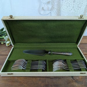 Puede incluir: Un estuche de cubiertos vintage rectangular abierto, con exterior crema e interior de fieltro verde. Contiene una espátula plateada para tartas y dos filas de tenedores y cucharas plateadas. El estuche está sobre una superficie de madera.