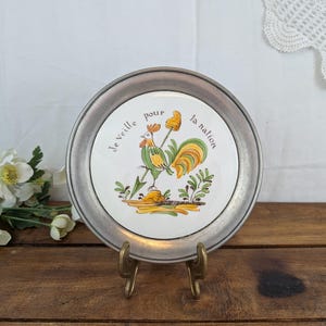 May include: A round decorative plate with a metallic pewter-toned rim and a white ceramic centre. The centre features a colourful illustration of a rooster and the French text Je veille pour la nation, surrounded by green and yellow foliage accents.