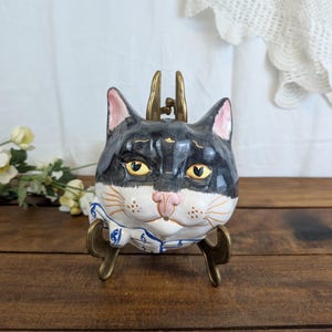 Peut inclure: Figurine en céramique en forme de tête de chat avec fourrure noire et blanche, oreilles roses et yeux jaunes. Le chat porte un col à motifs bleus et blancs et est exposé sur un support en métal doré.