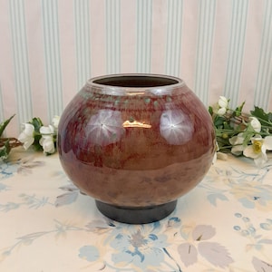 Puede incluir: Un jarrón redondo de cerámica marrón con un esmalte moteado y una base marrón oscuro. El jarrón tiene un color moteado marrón rojizo con toques de verde y blanco.