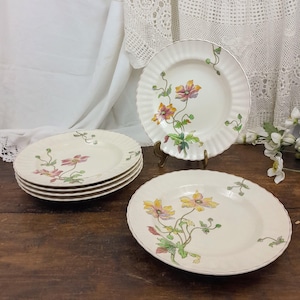 May include: Set of six white ceramic dinner plates with a floral design featuring pink, yellow, and green flowers. The plates have a slightly raised rim and a scalloped edge.