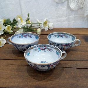 Peut inclure: Trois tasses à thé anciennes avec des anses, présentant un motif floral et architectural détaillé en bleu, rouge et vert. Les tasses sont blanches avec une bordure décorative, disposées sur une surface en bois foncé.