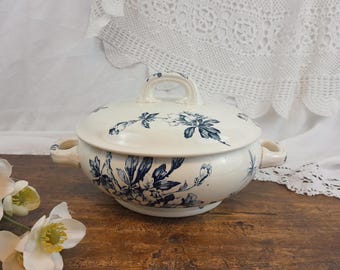 Op de afbeelding: Crèmekleurige keramische serveerschaal met een deksel, versierd met blauwe bloempatronen. De schaal heeft twee zijhandvatten en een handvat op het deksel. De schaal staat op een houten oppervlak.