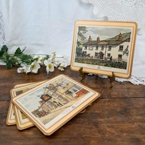Peut inclure: Ensemble de quatre sets de table vintage illustrant des pubs anglais. Chaque set de table présente une scène de pub différente avec le nom du pub imprimé en bas. Les sets de table sont beiges avec une bordure dorée.
