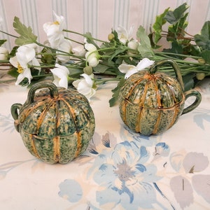 Op de afbeelding: Twee groene en bruine keramische pompoenvormige bekers met deksel en handvatten. De bekers staan op een witte tafel met bloemenmotief.