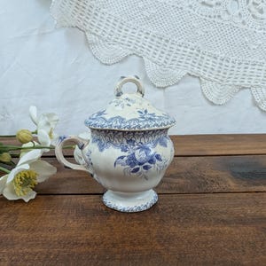 Peut inclure: Un sucrier en céramique blanc et bleu avec un couvercle, orné d'un motif floral. Le sucrier a une anse et repose sur un petit socle. Le couvercle a une petite poignée en forme de boucle. Le motif est bleu sur fond blanc.