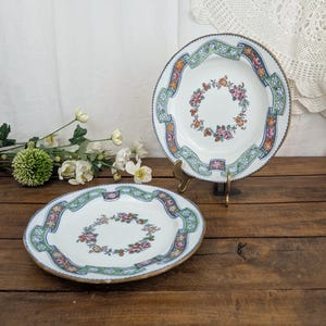 May include: Two white ceramic plates featuring a central floral wreath motif and a decorative border of green and orange ribbons with small flowers. The plates have a gold-toned rim and are displayed on a dark wooden surface with a metal stand.