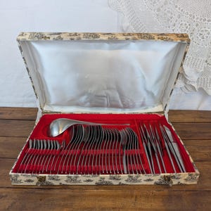 Peut inclure: Un service de couverts vintage dans un coffret décoratif. Le coffret a un extérieur crème avec un motif noir et blanc et un intérieur en velours rouge. L'ensemble comprend des cuillères, des fourchettes, des couteaux et une louche.