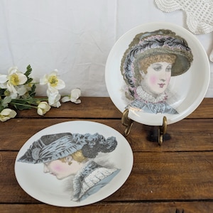 Peut inclure: Deux assiettes en céramique blanche ornées d'illustrations détaillées de personnes portant des chapeaux historiques et des cols à volants. Une assiette est posée sur un petit support en métal doré, l'autre est à plat, présentant des portraits vintage.