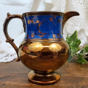 Op de afbeelding: Een blauwe en koperen kan met een gouden bloemenmotief. De kan heeft een rond lichaam en een uitlopende tuit. Hij heeft een handvat met een decoratief ontwerp.