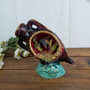 May include: A decorative ceramic vase in a dark burgundy colour with gold trim. The vase features a detailed interior scene with fish and aquatic plants. The base is turquoise with the text "Grau d'Agde".