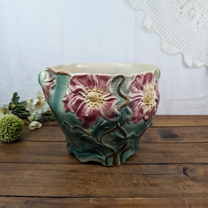 May include: A ceramic planter featuring a textured green base with raised, relief-style pink flowers and brown vine accents. The vessel has a scalloped rim and a wide, rounded body, showcasing a detailed botanical design with a glossy finish.