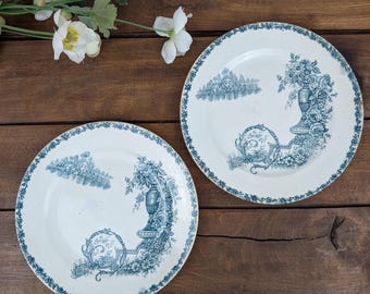 May include: Two white ceramic plates with a blue floral design. Each plate features a detailed blue illustration of a vase with flowers and a landscape scene. The plates have a delicate blue floral border.