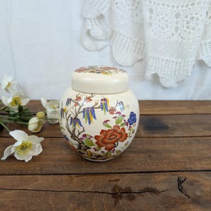 Peut inclure: Un pot en céramique crème avec un couvercle, orné d'un motif floral et d'oiseaux en rouge, bleu et jaune. Le pot est posé sur une surface en bois, avec des fleurs blanches et un rideau en dentelle en arrière-plan. Le pot mesure environ 15 cm de haut.