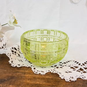 May include: A small, clear, green glass bowl with a square pattern. The bowl is sitting on a white lace doily.