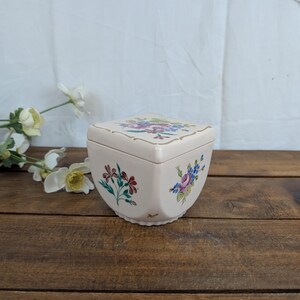 Puede incluir: Una caja de cerámica de color crema con tapa a juego, decorada con motivos florales pintados a mano en tonos rosa, azul, verde y marrón. El recipiente cuadrado tiene esquinas redondeadas, una base festoneada y detalles en tono dorado.