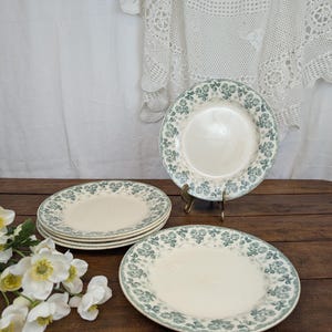 Op de afbeelding: Een verzameling vintage crèmekleurige borden met een delicaat groen bloemmotief rond de randen. De borden zijn gerangschikt op een donker houten oppervlak, sommige gestapeld en één op een messing standaard. Een wit kanten tafelkleed is op de achtergrond.
