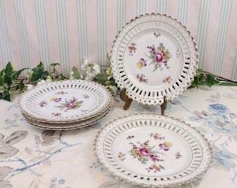 May include: Set of five white porcelain plates with a delicate floral design and a lace-like edge. The plates are stacked on top of each other, with the top plate slightly tilted to the side.