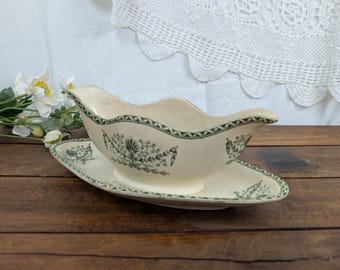 May include: Cream-coloured gravy boat and matching oval underplate. The set features a green floral and geometric pattern around the edges. The gravy boat has a curved handle and a spout. The set is on a wooden surface.
