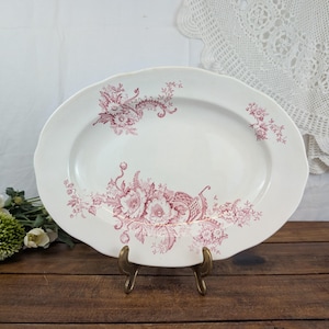 Könnte beinhalten: Eine ovale Servierplatte aus cremeweißer Keramik mit einem dunkelroten floralen Transferdruckmuster. Das Design zeigt detaillierte botanische Illustrationen von Blumen und Blättern, die entlang des Randes und in der Mitte angeordnet sind, mit gewelltem Rand.