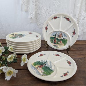 Puede incluir: Un juego de platos divididos vintage de color crema. Cada plato presenta una imagen circular central de un paisaje escénico y está decorado con detalles florales rojos y azules. Los platos están apilados y expuestos sobre una superficie de madera.