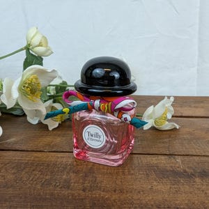 以下が含まれることがあります： 黒いキャップとカラフルなスカーフが首に巻かれた、ピンク色の正方形の香水瓶。「Twilly d'Hermès」とラベルされています。 黄色の中心を持つ白い花が、木製の表面にあるボトルの周りに配置されています。