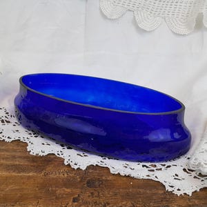 May include: A cobalt blue glass bowl with a rounded oval shape. The bowl is sitting on a white lace tablecloth.