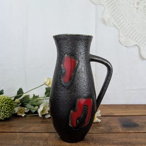 Puede incluir: Una jarra de cerámica negra con un acabado mate texturizado y dos parches abstractos rojos brillantes en la parte frontal. El recipiente presenta un borde acampanado, un asa curva en el lado derecho y una base cónica, mostrando un contraste claro.