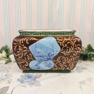 Peut inclure: Un pot rectangulaire brun avec des accents dorés et un motif floral bleu et blanc. Le pot a un bord vert avec un motif géométrique.