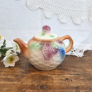 Può includere: Una teiera in ceramica con un motivo a intreccio di cesto, con un grappolo di uva viola e foglie verdi. La teiera ha un manico e un beccuccio marroni e un coperchio con un pomello rosso. La tavolozza dei colori è morbida e pastello.
