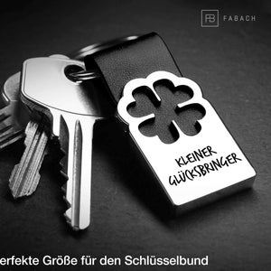 Könnte beinhalten: Ein silberner Schlüsselanhänger mit einem vierblättrigen Kleeblatt-Design und dem Text "KLEINER GLÜCKSBRINGER", was "kleiner Glücksbringer" bedeutet.