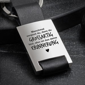 Könnte beinhalten: Ein schwarzer Leder-Schlüsselanhänger mit einem silbernen Anhänger, auf dem "Manchmal vergisst du, dass du GROSSARTIG bist, also ist das deine ERINNERUNG" mit einem kleinen Herzsymbol steht. Der Text "Liebevolle Worte als ständiger Begleiter" und "Lovely words as daily companion" ist unter dem Schlüsselanhänger gedruckt.