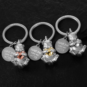 Könnte beinhalten: Drei silberne Schlüsselanhänger mit einem silbernen Otter-Anhänger, der ein Herz hält. Jeder Schlüsselanhänger hat ein rundes Schild mit dem Text "Ich liebe dich wie keinen anderen" darauf. Die Herzen sind gold, roségold und silber.