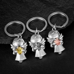 Könnte beinhalten: Drei silberne Engel-Schlüsselanhänger mit Flügeln, jeder mit einer andersfarbigen Blume. Die Blumen sind gold, roségold und silber.