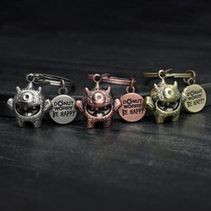 Könnte beinhalten: Vier Schlüsselanhänger mit einem Cartoon-Monster mit einem Auge und den Worten "Donut Worry Be Happy" auf einem runden Anhänger. Die Schlüsselanhänger sind in Silber, Kupfer, Gold und Bronze.