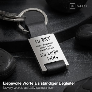 Könnte beinhalten: Ein silberner Schlüsselanhänger mit einem schwarzen Lederband. Der Schlüsselanhänger hat ein silbernes Rechteck mit dem Text "DU BIST mein Zuhause, mein Halt, mein Leben. ICH LIEBE DICH" eingraviert. Der Text ist in Deutsch und bedeutet "Du bist mein Zuhause, mein Halt, mein Leben. Ich liebe dich."